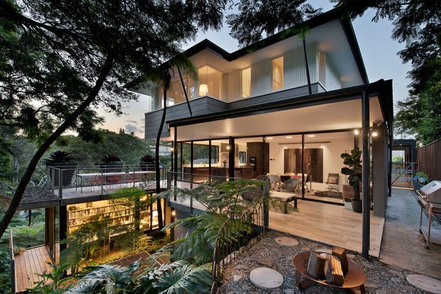 Twilight exterior view of a contemporary multi-level home set among mature trees and lush planting. The house features dark metal framing, expansive floor-to-ceiling glazing, projecting balconies, and a partially open lower-level living area with warm interior lighting. A landscaped courtyard with gravel, stepping stones, outdoor seating, and a fire pit occupies the foreground, while a lower illuminated level is visible through the garden.
