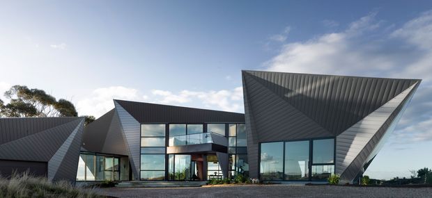 Exterior view of a striking contemporary multi-wing residence composed of sharply folded, angular roof forms and dark ribbed metal cladding. Large floor-to-ceiling glazed walls reveal a bright interior, while the building sits on a broad paved or gravel forecourt with sparse landscaping. The sculptural roof geometry dominates the composition.