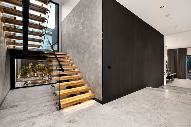 Contemporary open-plan entry hall featuring a floating timber staircase with glass balustrades, a dramatic black vertical timber feature wall, and a textured grey concrete-like wall. The stair void has a black structural element and skylit glazing, with a planted courtyard visible beneath and a dining area beyond the hall. The palette is monochromatic and refined, balanced by warm timber stair treads and natural greenery.