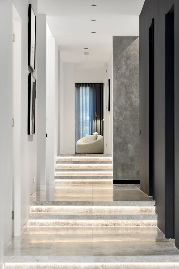 A refined multi-level interior hallway with broad pale stone stair treads illuminated by integrated LED strip lighting. White walls and concealed cabinetry contrast with a dark vertical-panelled wall and a large textured grey feature surface. Recessed ceiling lights lead toward a softly furnished sitting area with full-height dark sheer curtains at the far end. The space has a clean, luxurious and highly architectural appearance.