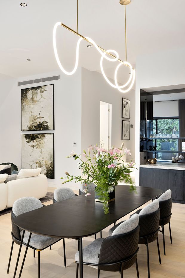 Bright contemporary open-plan dining and living space with a large dark oval dining table surrounded by upholstered chairs, a sculptural looping pendant light, pale timber flooring, white walls, abstract artwork, and a dark built-in kitchen visible at right. A floral centrepiece adds colour to the otherwise neutral interior.