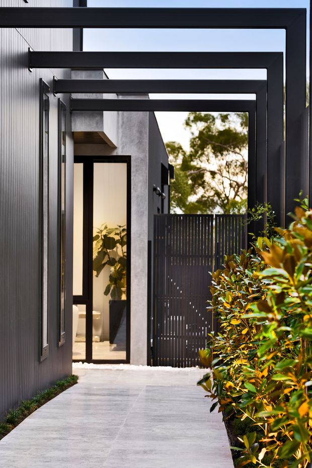 A narrow landscaped entry walkway leads toward a modern home entrance. The facade features dark vertically ribbed cladding, exposed concrete, black-framed glazed doors, and a series of bold rectangular overhead beams forming a covered passage. A vertical slatted privacy gate and dense shrubs frame the path, with a softly lit interior visible through the glass.