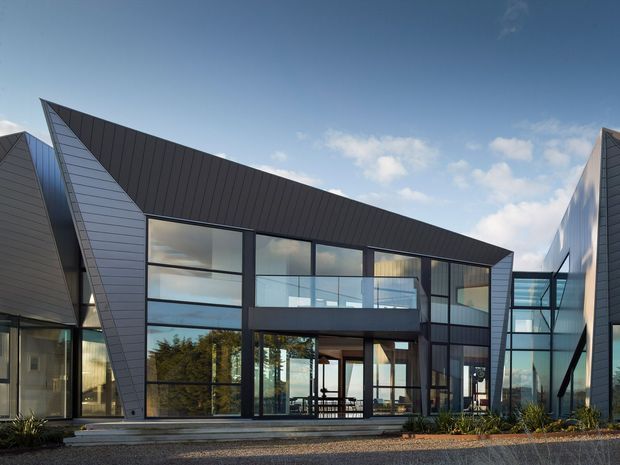 Exterior view of a contemporary angular building composed of dark standing-seam metal roof planes, grey cladding, and extensive floor-to-ceiling glazing. The central facade features a recessed upper balcony and glazed entrance, with connected wings creating a strong geometric composition. Low landscaping and a paved forecourt sit in front under a clear sky.