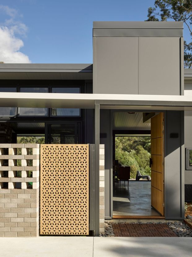 Front exterior view of a contemporary single-storey home entrance. The facade combines dark glazing, grey smooth cladding, a broad projecting entry canopy, a timber pivot door, and patterned breeze-block screens set into a low masonry wall. The entry opens through to a bright interior and landscaped greenery beyond.