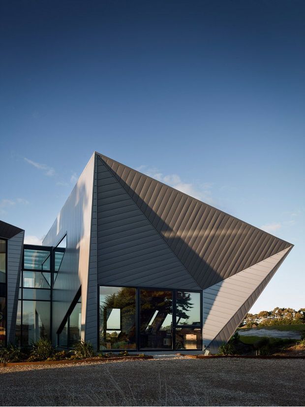 Exterior architectural photograph of a highly geometric contemporary building with dramatically folded, angular roof and facade planes. Dark vertically ribbed metal cladding forms the central triangular volume, while lighter metal panels wrap the projecting side plane. Large black-framed glazed openings reveal a small amount of interior space and reflected trees. The building sits in an open landscaped setting under a clear blue sky.