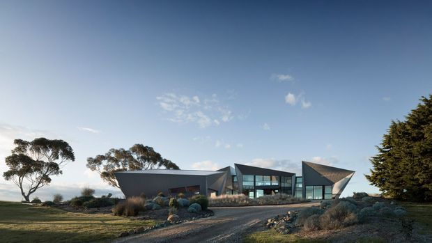 Wide exterior view of a low, contemporary architectural residence set in a landscaped rural or coastal-looking site. The building is composed of angular geometric volumes with dark grey cladding, extensive glazing, pitched and folded roof forms, and a gravel driveway leading toward the entrance. Established trees, ornamental grasses, and rounded shrubs surround the house under a clear blue sky.