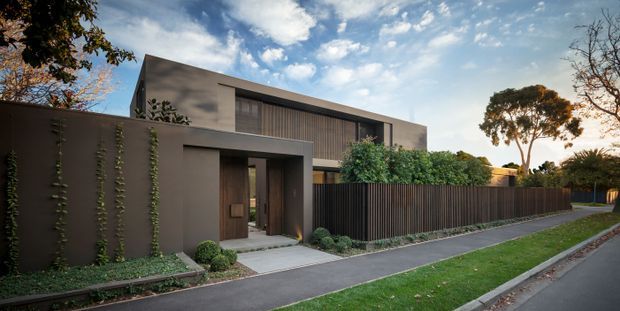 Street-level exterior view of a contemporary two-storey residence with a restrained flat-roofed form, smooth dark grey rendered walls, vertical timber-look screening and a recessed covered entry. A tall privacy fence, landscaped planting, lawn verge and paved pedestrian path frame the facade.