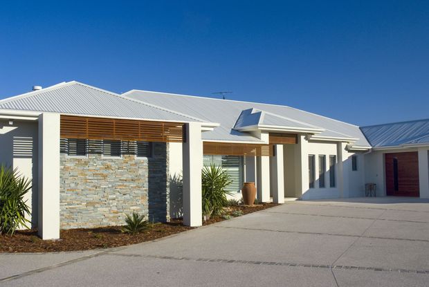 Single-storey contemporary residential building viewed from the driveway, with white rendered walls, pale grey metal pitched roofs, broad roof overhangs, timber louvres and screens, and a prominent stacked-stone feature wall. The paved forecourt leads toward an attached garage or covered parking area. Sparse drought-tolerant planting borders the facade under a clear blue sky.