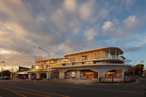 Exterior street-level view of a contemporary multi-storey commercial building at dusk, featuring sweeping curved floor plates, extensive glazing, projecting balconies, a rounded roofline, and a covered pedestrian frontage with retail or hospitality spaces at ground level. The building occupies a prominent urban corner site with surrounding streetscape and mature trees.