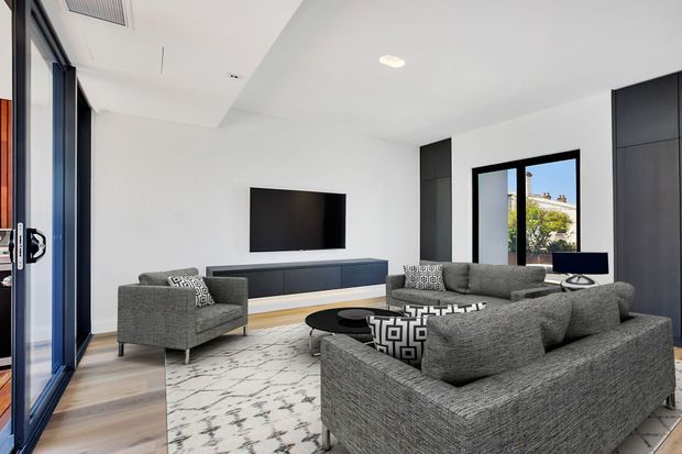 Contemporary living room with a large grey sectional sofa, matching armchair, black oval coffee table, patterned area rug, wall-mounted television, and extensive dark built-in cabinetry. White walls and ceiling, pale timber flooring, black-framed glazing, and a glazed door create a bright, refined interior.