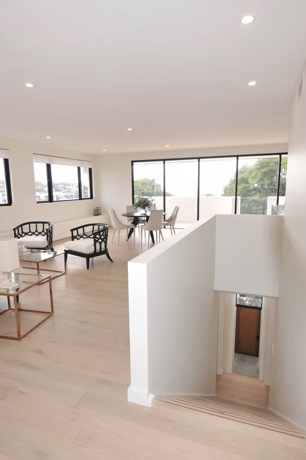 Bright, minimalist open-plan upper-level living and dining area with pale timber flooring, white walls and ceiling, recessed downlights, and expansive black-framed windows. A stairwell with white walls descends to a lower level, while the room contains contemporary dining furniture, black-framed lounge chairs, and a glass coffee table.