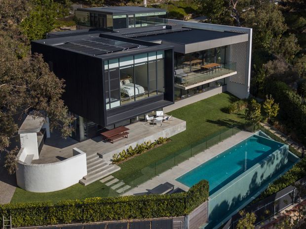 Aerial view of a large contemporary multi-level residence set within a landscaped suburban property. The dark, flat-roofed house features extensive floor-to-ceiling glazing, projecting volumes, rooftop elements, a perforated screen wall, balconies, terraces, and a long rectangular swimming pool. Outdoor entertaining areas, lawns, hedges, paving, and curved rendered walls surround the home.