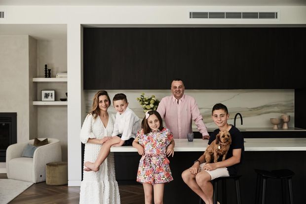 A bright, contemporary open-plan kitchen and living area photographed with a family gathered around a large island. The kitchen features dark full-height cabinetry, a pale stone or marble-look splashback, a light island benchtop, black stools, and integrated appliances. A neutral living area with built-in shelving and upholstered seating is visible to the left.