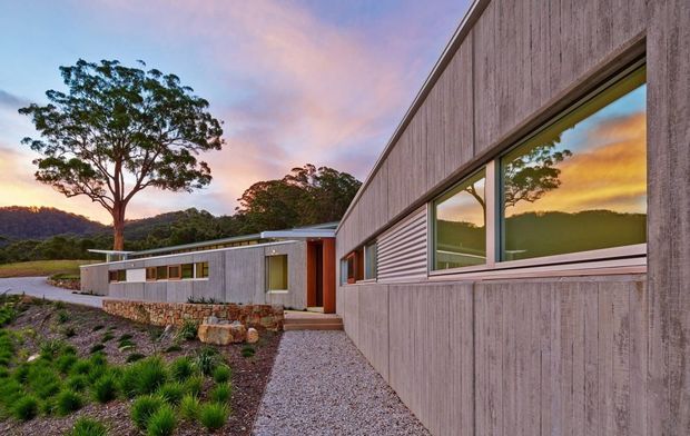 Exterior architectural view of a low-profile contemporary house at sunset. The composition focuses on a long, angular facade clad in vertically textured grey material, with broad horizontal windows reflecting the warm sky and surrounding landscape. A gravel path leads past a restrained landscaped garden toward a recessed timber-toned entry, while a connected lower wing and stone retaining wall extend into the site. The setting appears rural or bush-fringed, with hills and mature trees beyond the property.