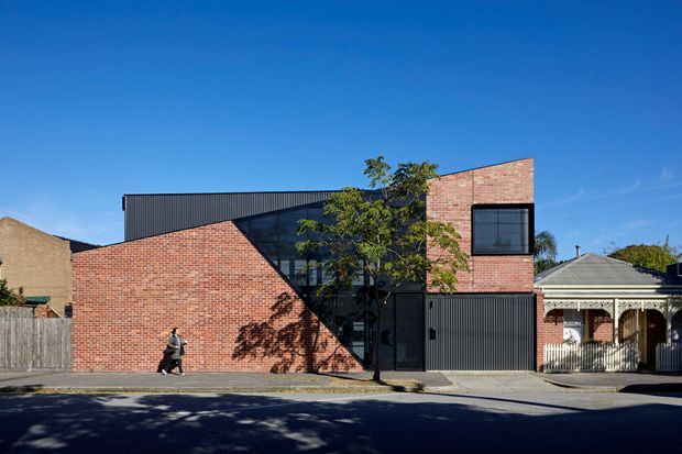 Street-facing view of a contemporary multi-level brick building with a strong geometric facade. Red brick walls are combined with dark ribbed metal cladding, large dark-framed glazing, and a prominent diagonal glazed section. A traditional neighbouring house is visible at the right edge, with mature street trees and a clear blue sky.