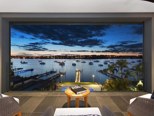 A refined waterfront living area or lounge opens onto a dramatic floor-to-ceiling glazed view of a marina at dusk. The interior features large-format dark grey tiled flooring, slim dark window framing, and woven lounge chairs arranged around a small round timber table. A private jetty and boats are visible across the water, with warm exterior lighting contrasting against the blue evening sky.
