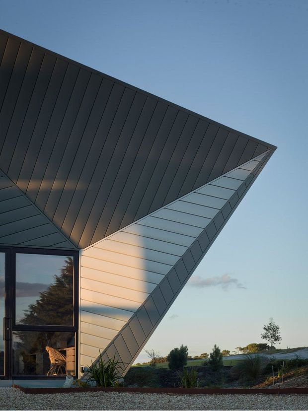 Dramatic close-up exterior view of a contemporary angular building with a sharply folded, wing-like roof and dark standing-seam metal cladding. A tall glazed doorway reflects surrounding trees and reveals a chair inside. The building sits in a landscaped, low-vegetation setting beneath a clear evening sky, with warm sunlight accenting the pale metal wall panels.
