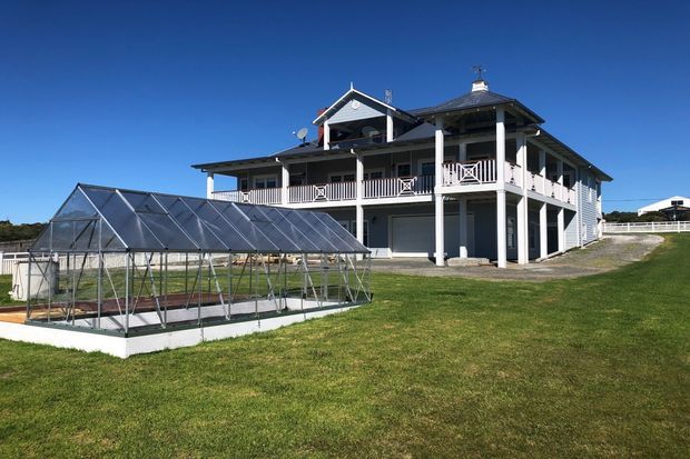 Exterior view of a large elevated multi-storey home with a wraparound covered veranda, white railings, dark roofing, and an attached greenhouse in the foreground. The property sits in an open grassy setting under a clear blue sky.