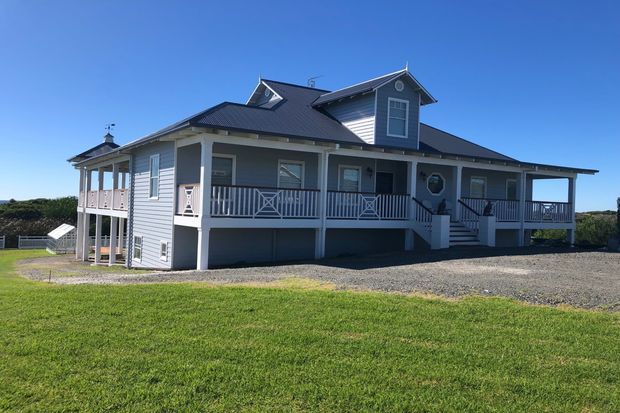 Exterior view of a raised, single-storey coastal-style house with light blue-grey weatherboard cladding, a dark pitched metal roof with dormer windows, broad covered verandas, white posts and decorative railings, and a gravel driveway. The building sits in an open grassy setting under a clear blue sky.