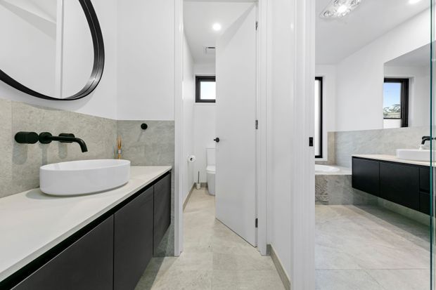 Bright contemporary bathroom with a black floating vanity, white vessel basin, dark wall-mounted tapware, pale stone-look wall and floor finishes, and a large round mirror. A separate bathing area with a freestanding bathtub and matching dark vanity is visible through the central doorway and reflection. White doors, black-framed windows, and recessed lighting create a clean, high-contrast scheme.