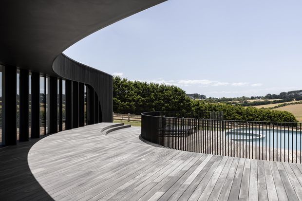 A dramatic contemporary curved building facade opens onto a broad timber deck overlooking a rural landscape. The deck follows the sweeping geometry of the dark-clad structure and is enclosed by slim black metal balustrading, with a swimming pool visible beyond. Floor-to-ceiling glazing lines the curved facade, creating a strong indoor-outdoor connection.
