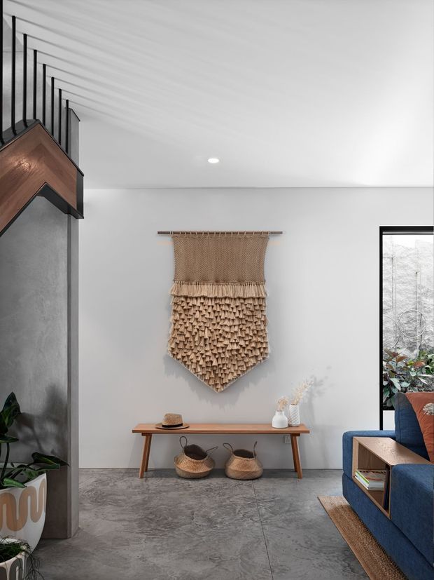 A modern open-plan living area with a pale grey plaster wall, polished grey concrete-look flooring, and black-framed glazing. A sculptural woven wall hanging forms the focal point above a slim timber console table, accompanied by woven baskets and simple decorative objects. A blue upholstered sofa and timber side table are partially visible at right, while a dark textured partition and warm timber architectural element frame the left side. The space has a restrained, earthy palette with contemporary styling and natural woven accents.