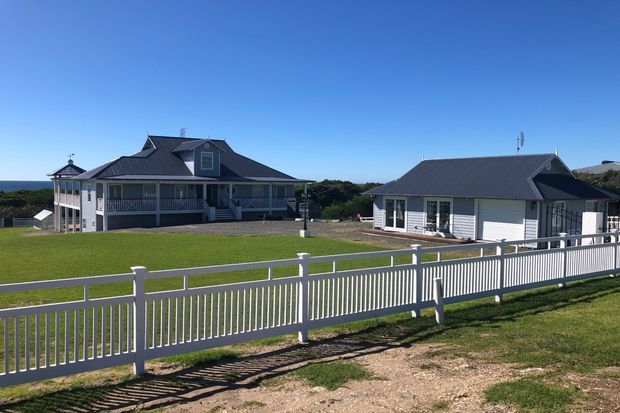 A spacious coastal residential property viewed from outside, featuring a large light-coloured multi-level house with covered verandas and a steep dark pitched roof, alongside a separate single-storey garage or outbuilding. A white picket-style fence encloses the neatly landscaped lawn, with open ocean visible in the distance under a clear blue sky.