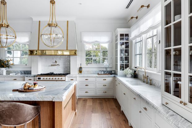 Bright, refined kitchen with white shaker-style cabinetry, marble-look benchtops and splashback, warm timber flooring, brass hardware and fixtures, a large island, rangehood, and abundant multi-pane windows. Gold globe pendant lights and glass-front cabinetry add a classic decorative touch.
