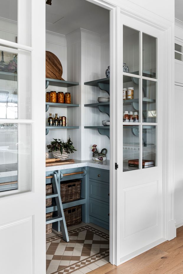 A compact, beautifully detailed walk-in pantry with white vertical wall panelling, deep blue-grey built-in cabinetry and open shelving, glass-panelled white doors, patterned geometric floor tiles, wicker storage baskets, and neatly arranged pantry goods and serving pieces. The space has a refined traditional character with strong contrast between the pale envelope and dark joinery.