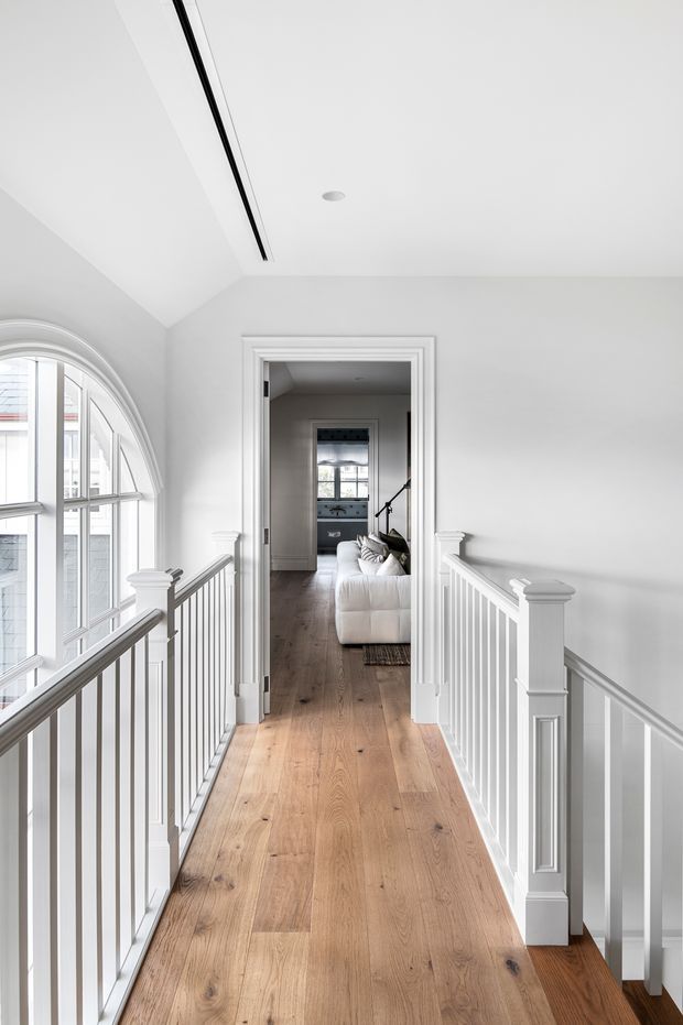 Bright, refined upper-level hallway or landing with white painted walls, detailed traditional trim, white balustrades, and a warm timber floor. A large arched window floods the space with natural light, while an open doorway leads toward a living room with a white sofa and visible staircase beyond.