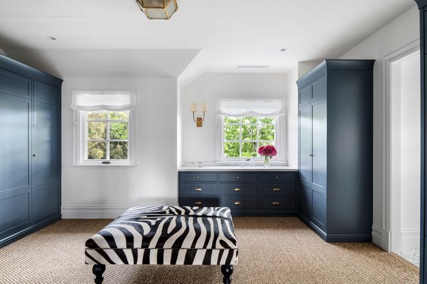 Bright, tailored bedroom with deep navy built-in wardrobes and a matching low dresser beneath two sash windows. The room has white walls and ceiling, white traditional trim, natural woven carpet, and a zebra-pattern upholstered bench at the foot of the bed. Brass-toned wall lights and a small decorative ceiling fixture add restrained detail.
