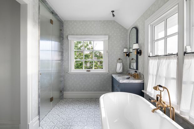 Bright, elegant bathroom featuring a freestanding white bathtub beneath large sash windows with café curtains, a blue-grey vanity with brass hardware and fittings, a frameless glass shower enclosure, patterned grey-and-white floor tiles, and subtly patterned pale grey wall covering. A small window and arched mirror add architectural character.