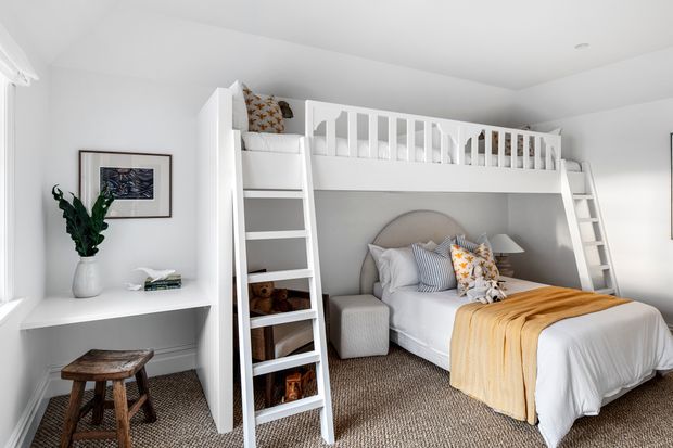 Bright, compact bedroom featuring a white built-in loft bunk structure with railings and ladders above a double bed. The room has white walls and joinery, a neutral textured carpet, warm mustard bedding, striped and patterned cushions, a small desk beneath the window, and a simple wooden stool. The overall design is clean, playful, and space-efficient.
