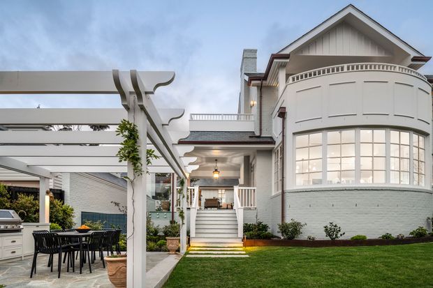 Exterior view of a refined white multi-level residence with a prominent curved, bay-like glazed facade, gabled roof, balcony railing, and light brick lower walls. A covered outdoor entertaining area with a pergola, barbecue, dining table, paving, landscaped lawn, and steps leading to the entry are visible.