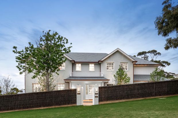 A large two-storey residential house viewed from across a neatly maintained lawn. The light rendered facade features a grey pitched roof with multiple gables, white-framed windows, a small upper balcony, and a central gated entry partly screened by a dark woven fence. Mature trees and landscaped greenery frame the property.