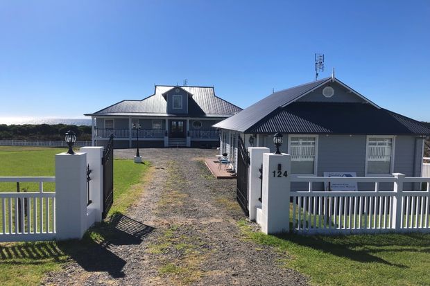 Front exterior view of a low-set residential property with two connected pitched-roof volumes, dark corrugated metal roofing, pale grey cladding, white rendered entrance pillars, and a white metal picket-style fence. A gravel driveway leads through the front gate toward the house, with open lawn on either side.