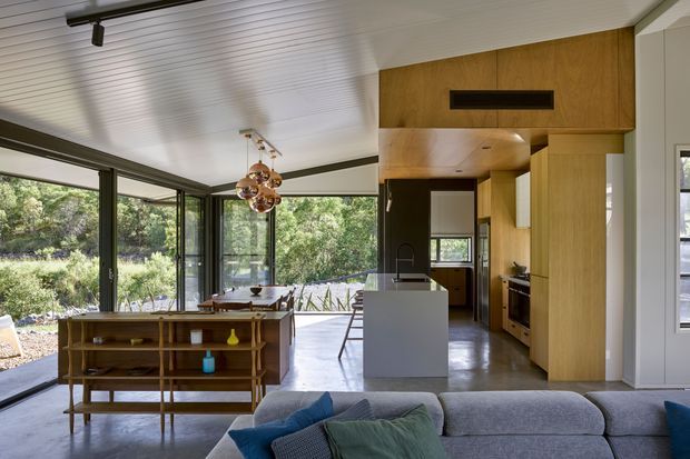 Open-plan contemporary living and kitchen interior with extensive floor-to-ceiling glazing overlooking greenery. The room features a white linear-panel ceiling, dark-framed sliding glass doors, polished concrete flooring, warm timber cabinetry and wall joinery, a central kitchen island, a dining area, and a large sectional sofa in the foreground.