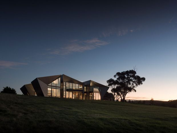 A contemporary, low-profile building set within a broad grassy landscape and photographed at dusk. The angular dark-clad facade features several sharply folded roof and wall planes, extensive floor-to-ceiling glazing, and warm interior lighting visible through the windows. A mature tree stands beside the building, with an open rural-looking horizon beyond.