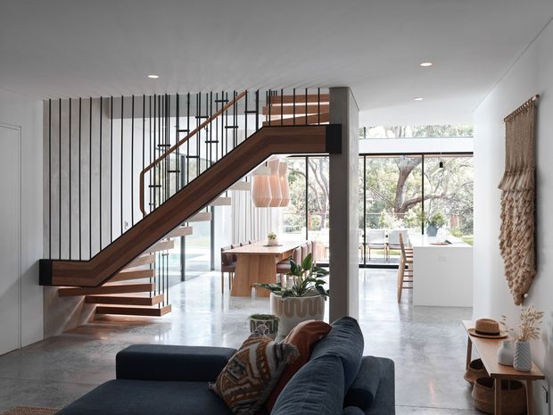 A bright, open-plan contemporary interior viewed from a living area, featuring a sculptural timber-and-black-metal staircase with open risers, a dining space beyond, polished concrete flooring, extensive glazing, and a dark sectional sofa in the foreground. Vertical black stair balusters and timber detailing create a strong architectural feature, while woven wall art and indoor plants add warmth.