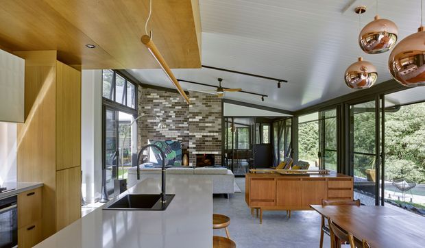 Open-plan contemporary kitchen, dining and living area with extensive floor-to-ceiling glazing. The kitchen features a white island with an integrated dark sink and tap, warm timber cabinetry, and a timber-clad ceiling section. A patterned brick feature wall anchors the living area, while a timber sideboard and dining table sit beside large windows overlooking greenery. Copper spherical pendant lights and black-framed glazing add contrast.