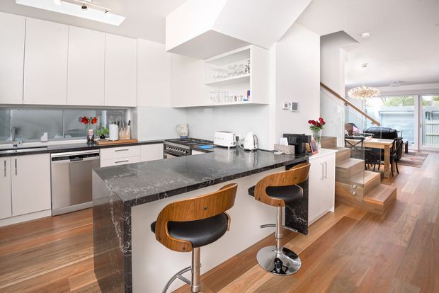 Bright open-plan kitchen with white flat-panel cabinetry, a dark stone island with waterfall ends, integrated appliances, timber flooring, and two curved timber-and-black bar stools. A dining area and stairway are visible beyond the kitchen, with a large rear window or glazed door bringing in natural light.