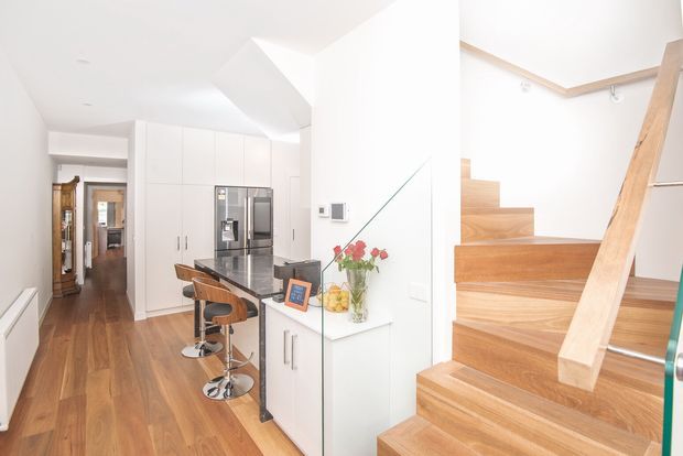 Bright contemporary open-plan kitchen and stair hall with white floor-to-ceiling cabinetry, stainless-steel appliances, a dark island with timber bar stools, and warm timber flooring. A timber staircase with a simple handrail rises at right, while a hallway extends toward the rear of the home.