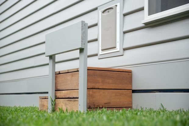Close-up exterior view of a light grey weatherboard-clad building, showing a white utility or service panel, a small window, a grey metal post-mounted box, and a timber planter or built-in box at ground level. Grass is visible in the foreground.