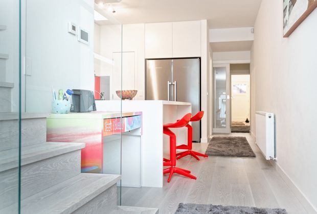 Bright contemporary open-plan kitchen and hallway viewed from a stairway. The space features white floor-to-ceiling cabinetry, a stainless-steel refrigerator, a white island with a colourful illuminated or reflective panel, vivid red bar stools, pale grey timber-look flooring, a glass stair balustrade, and a grey runner rug. A corridor and entry door extend beyond the kitchen.