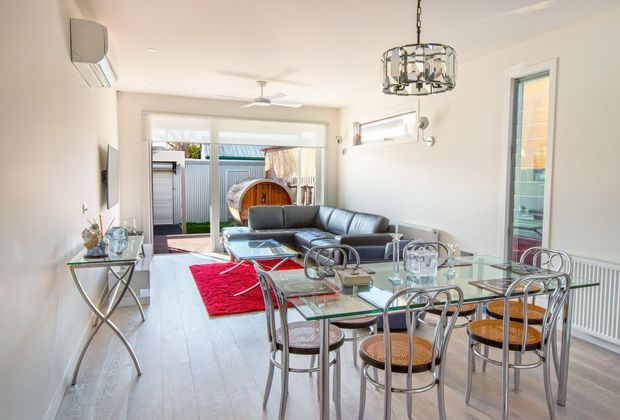 Bright open-plan interior combining a living room and dining area. The space features pale timber-look flooring, white walls, large glazed openings, a dark grey leather sectional sofa, a glass dining table with metal-framed chairs, a red rug, a contemporary pendant light, and a ceiling fan. Outdoor decking and a spa or tub are visible beyond the glazing.