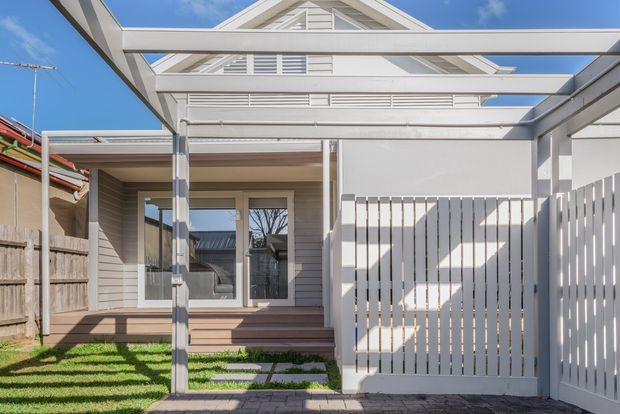 Bright exterior view of a compact, contemporary residential dwelling with a covered entry and carport-like overhead structure. The facade combines pale horizontal weatherboard cladding, white painted framing, a gabled roof with louvred ventilation panels, and a white vertical-slat privacy fence. A small grassed front yard, paved stepping stones, timber steps, and glazed entry doors are visible.
