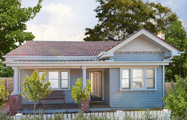 Front exterior view of a renovated single-storey house with a broad tiled pitched roof, pale blue horizontal weatherboard cladding, white decorative trims, a small covered entry porch supported by columns, and a gabled front projection with shingle detailing. Brick column bases, a low white picket fence, and mature trees surround the facade.