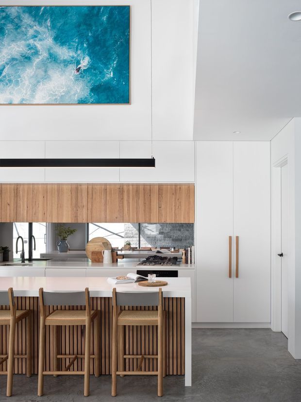A bright contemporary kitchen with a large white island and integrated seating for four. The island has a vertically slatted timber base and a white benchtop, while the rear wall combines warm timber overhead cabinetry with white cabinetry and a dark cooktop. Tall white pantry cabinetry with timber handles occupies the right side. The room features a smooth grey floor, white panelled walls and ceiling, a long black linear pendant, and a large turquoise ocean-inspired artwork.