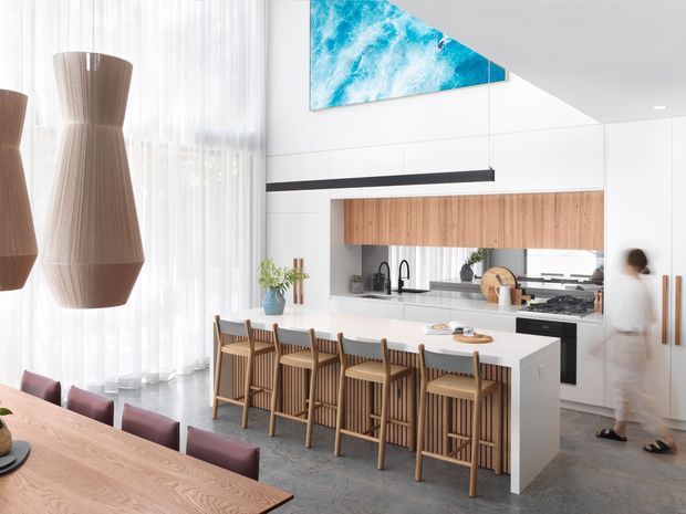 Bright contemporary open-plan kitchen and dining space with a large white island, timber bar stools, extensive white cabinetry, and a timber dining table in the foreground. Floor-to-ceiling sheer curtains bring in abundant natural light, while a sculptural timber pendant and vivid blue abstract feature artwork add visual interest. A partial upper-level void or high ceiling is visible above the kitchen.