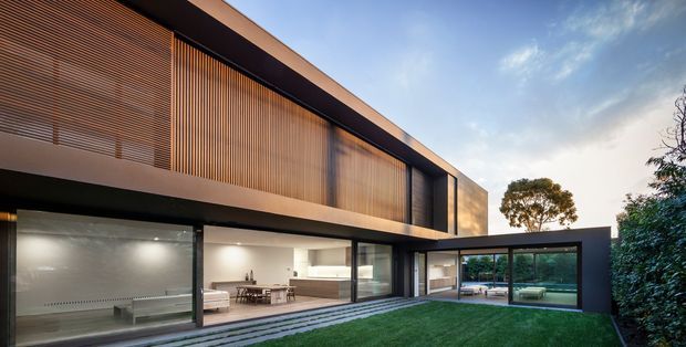 A contemporary two-storey residence viewed from the rear garden, with a long rectilinear form, dark framed glazing, warm timber vertical screening on the upper level, and expansive ground-floor windows opening toward a landscaped lawn and pool courtyard. The composition emphasises strong horizontal roof and floor lines, indoor-outdoor connection, and a restrained black, timber, and glass palette.